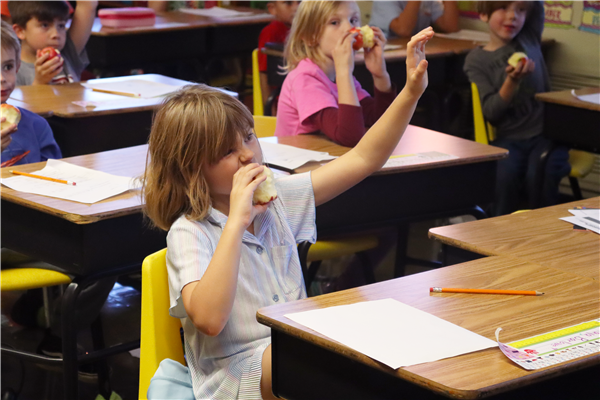 A girl sits at a desk biting an apple in her right hand and raising their left hand.